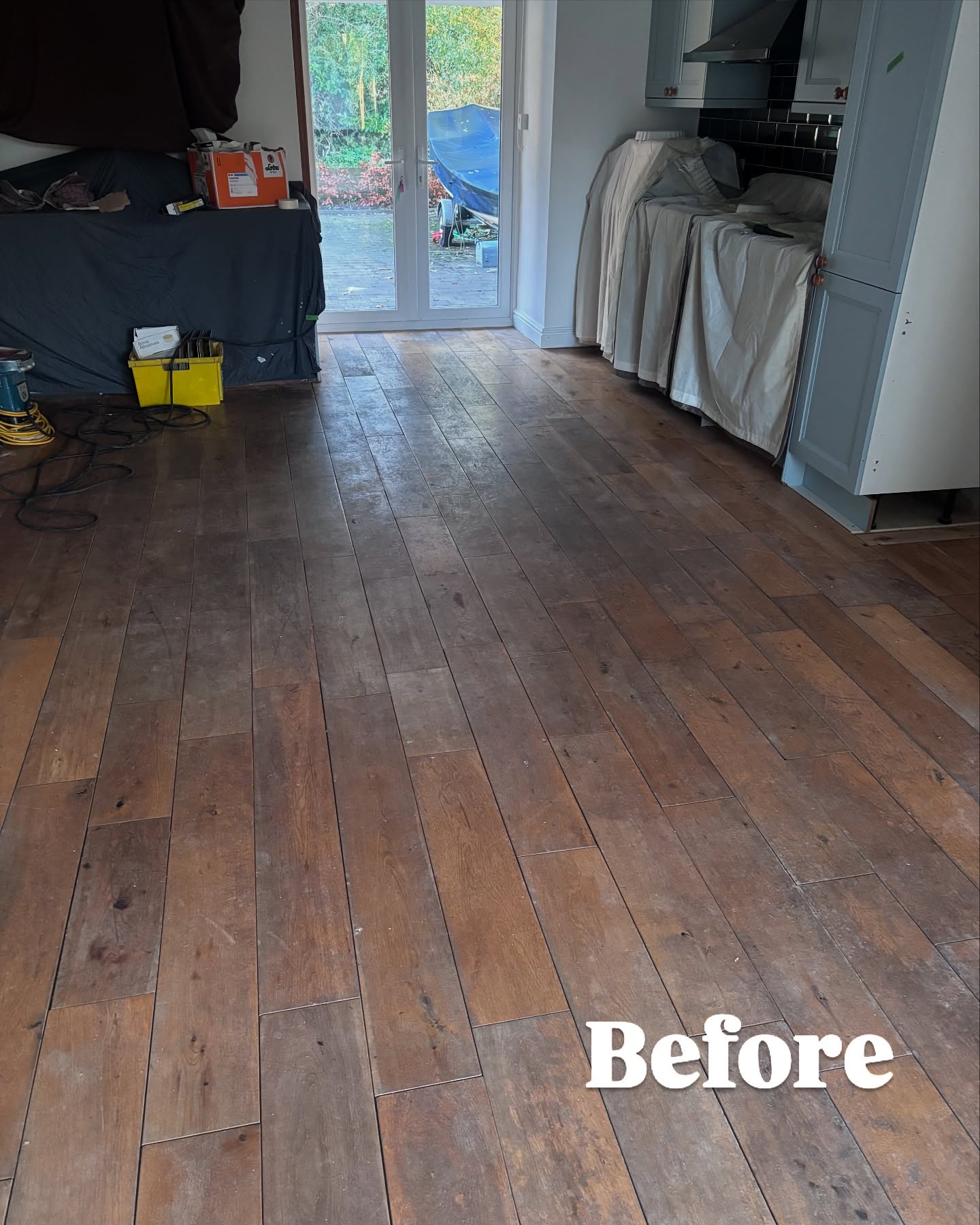 Worn dark hardwood floor in an open-plan kitchen-diner before restoration — tools and dust sheets visible mid-renovation